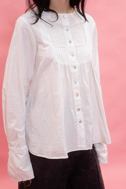 A white long-sleeve button-up blouse with a smocked chest panel, worn by a model against a solid pink background.