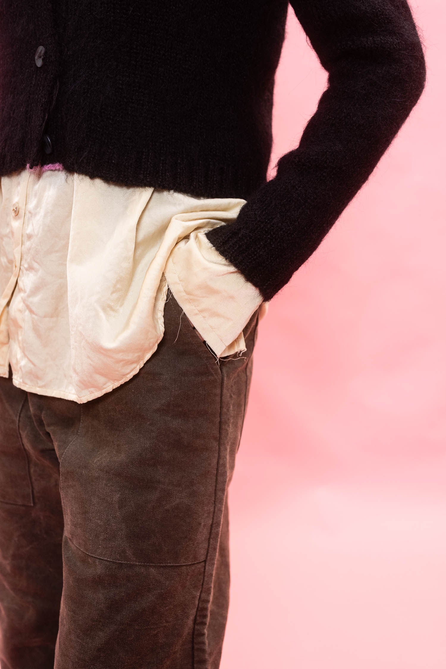 A close-up of a person wearing a black knit cardigan over a cream satin shirt and brown cotton trousers, standing against a solid pink background.