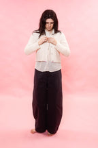 A woman stands barefoot against a solid pink studio background, wearing a cream-colored cropped cardigan over a white top and dark brown wide-leg pants.