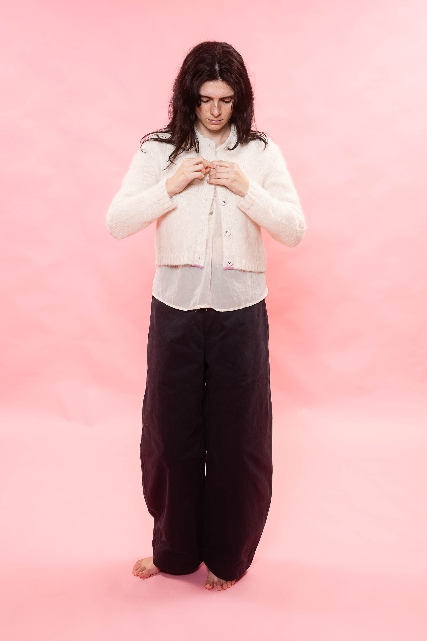 A woman stands barefoot against a solid pink studio background, wearing a cream-colored cropped cardigan over a white top and dark brown wide-leg pants.