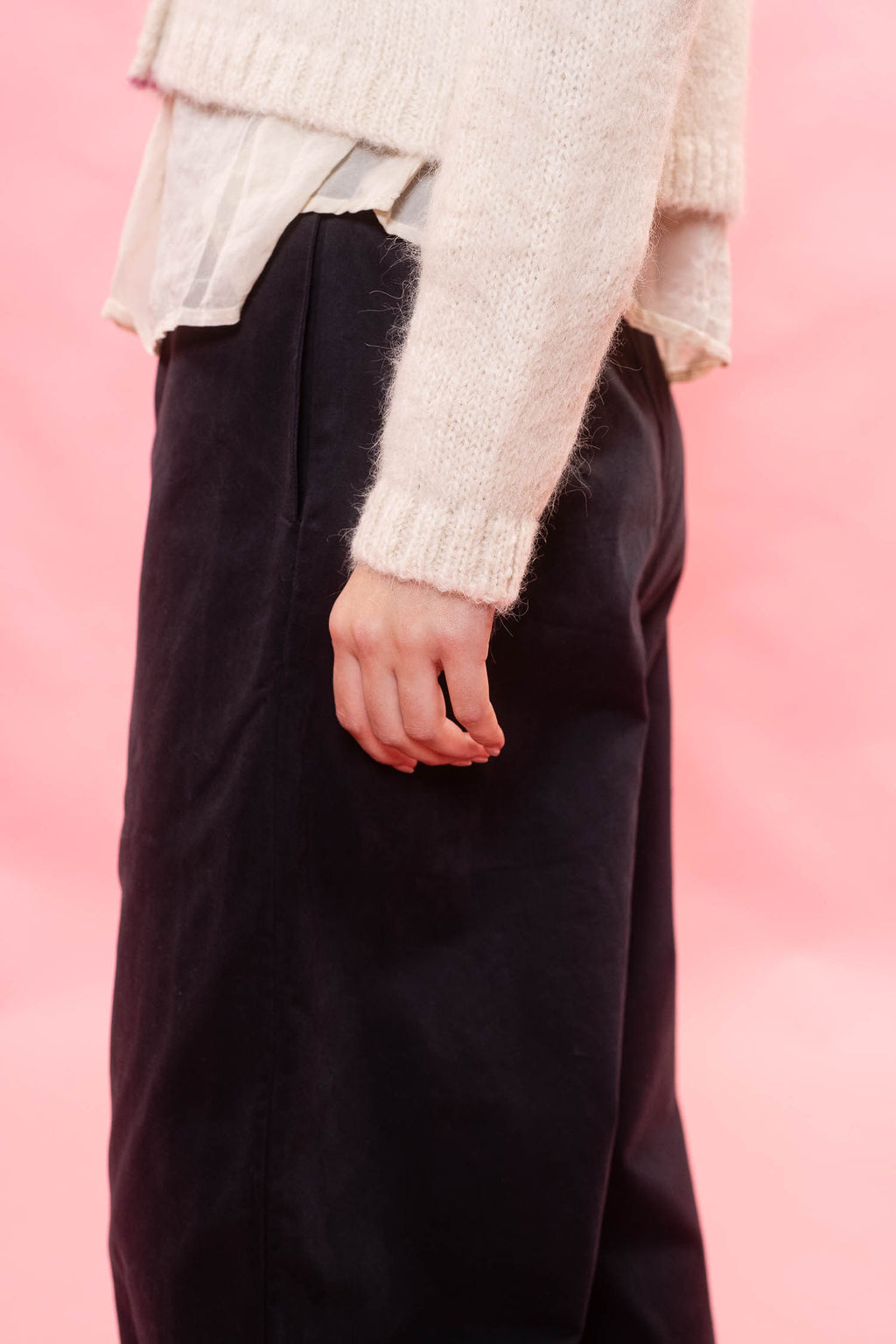 A close-up of a person wearing dark wide-leg trousers and a fluffy cream knit sweater against a solid pink background.
