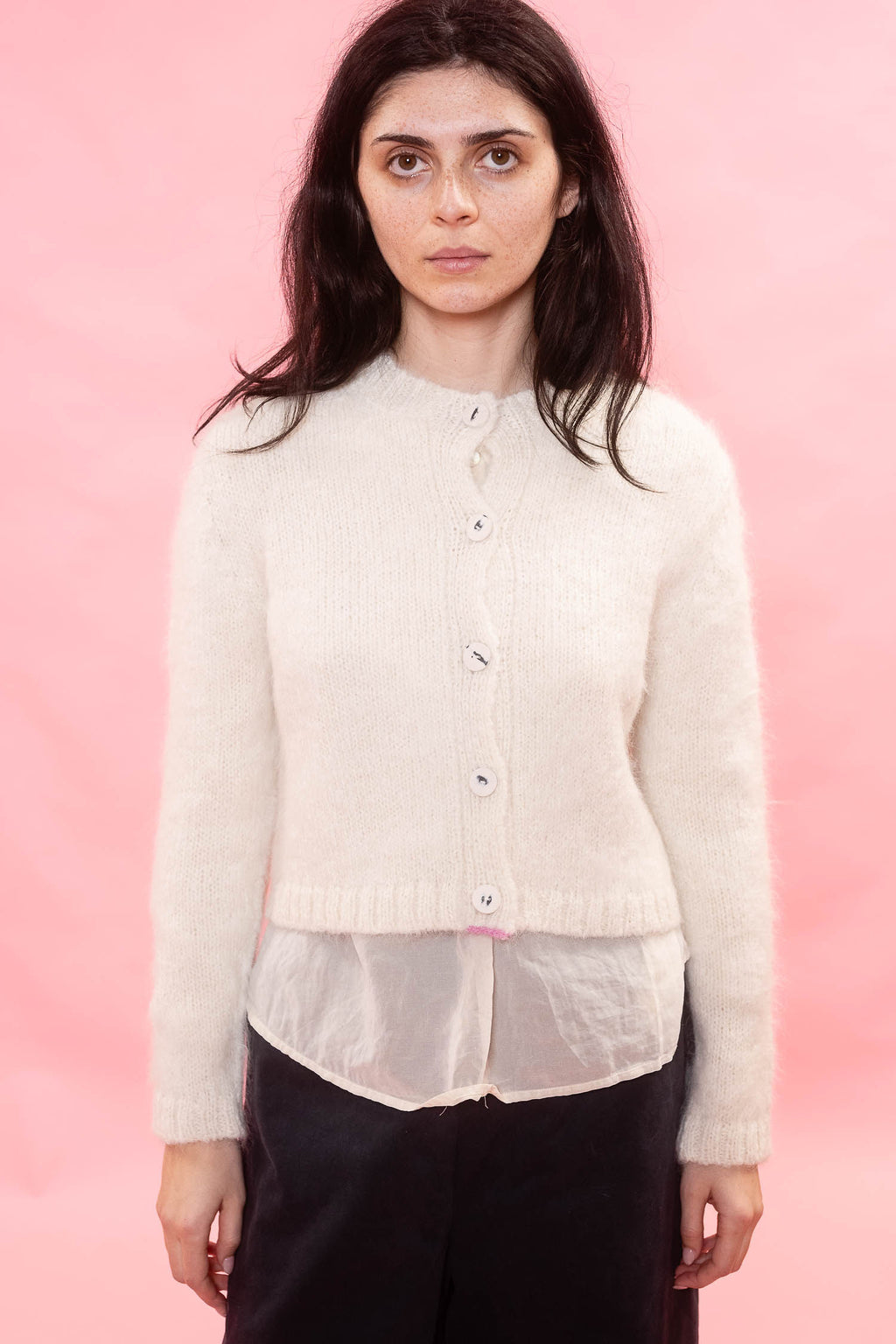 A woman modeling a cream-coloured, long-sleeve knit cardigan with a button-up front, layered over a sheer white shirt and black trousers, set against a solid pink background.