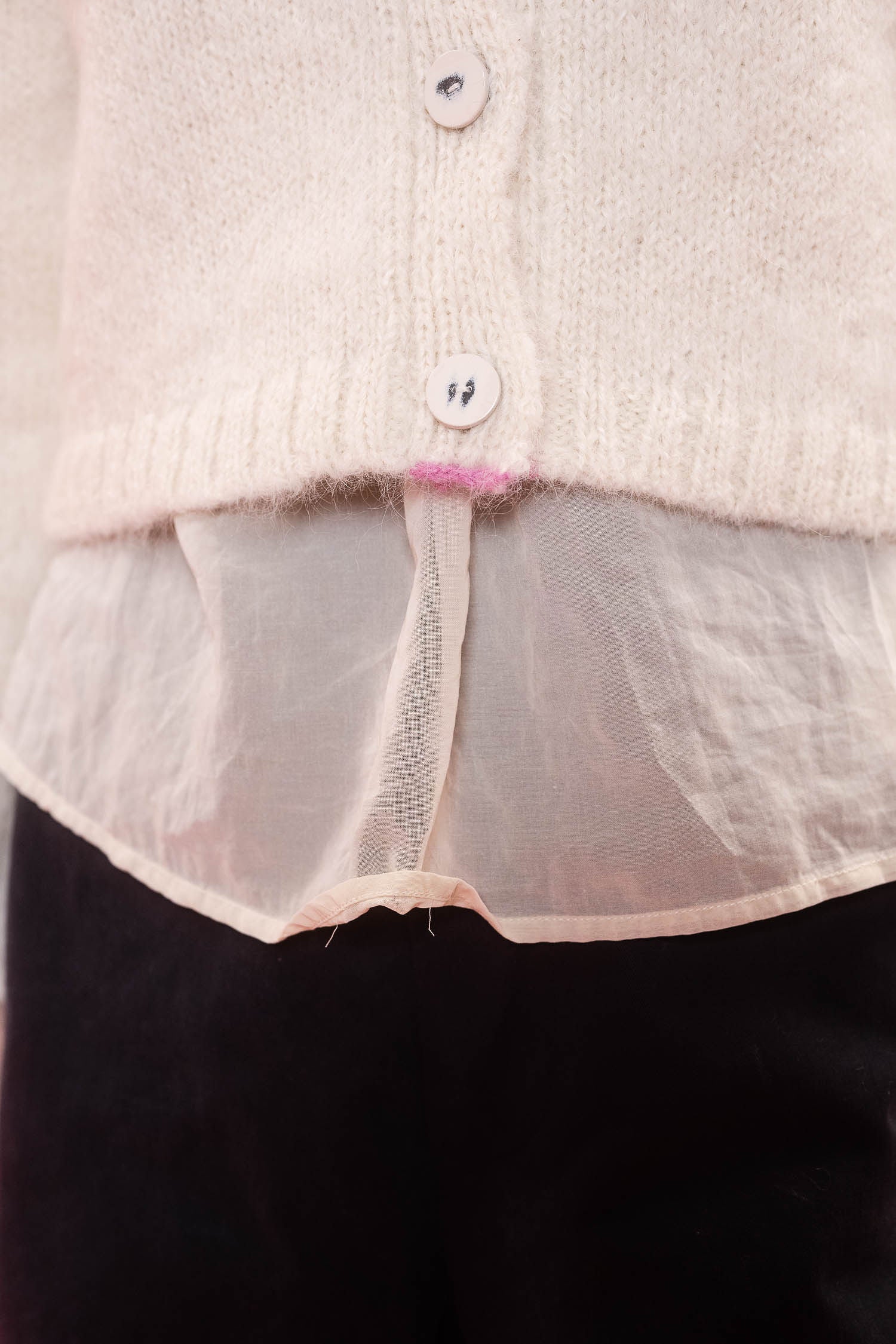 Close-up of a cream knit cardigan with white buttons and pink stitch detail at hem, layered over a sheer cream blouse, paired with black bottoms.