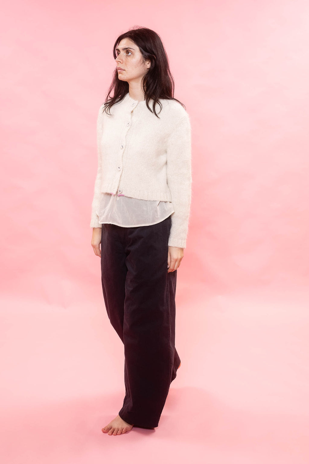 A woman stands barefoot against a solid pink studio backdrop, wearing a cream-colored cropped cardigan over a sheer white top and dark brown wide-leg trousers.