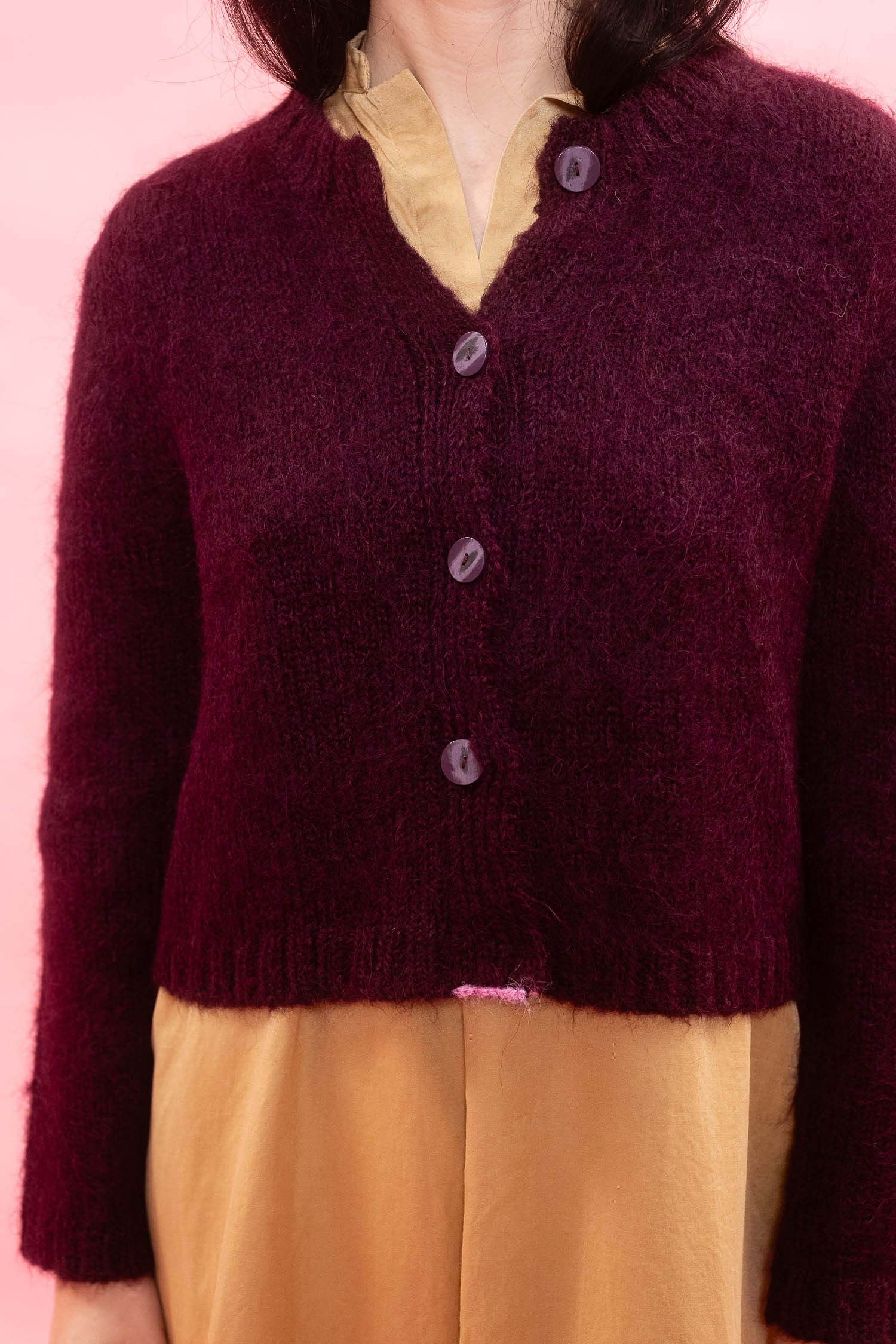 A close-up of a person wearing a fluffy knit alpaca cardigan in deep plum over a mustard yellow dress, set against a solid pink background.

