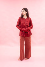 A woman stands barefoot against a solid pink studio backdrop, wearing a matching two-piece outfit consisting of a long-sleeve button-front top in a deep red-burgundy satin-like fabric and wide-leg pants in a rust red corduroy or textured fabric.