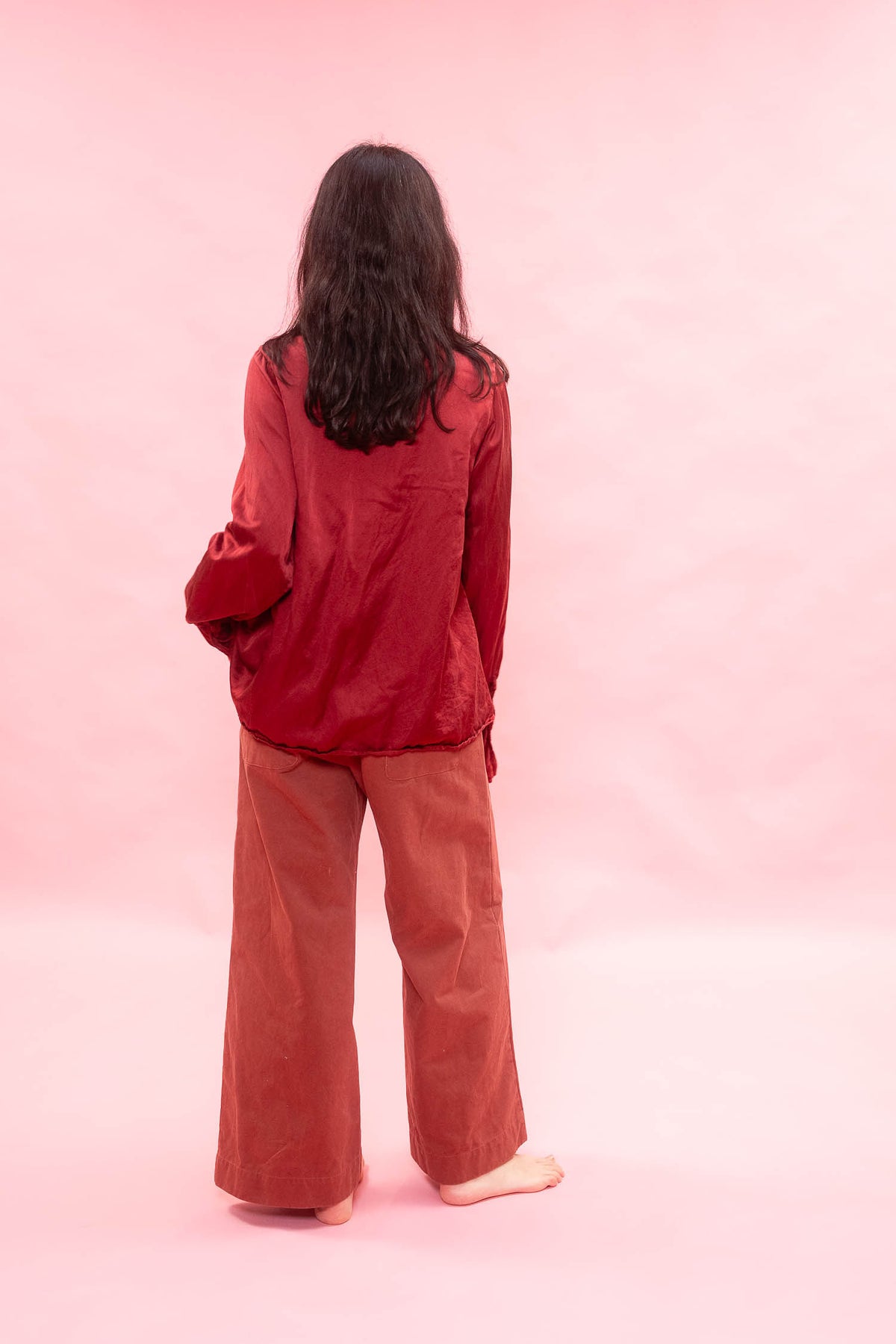 A person standing with their back to the camera, wearing a shiny dark red satin-like long-sleeve top and wide-leg rust-brown pants against a solid pink studio background.