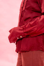 A close-up of a person wearing a long-sleeve red button-up shirt and matching red pants against a soft pink background.