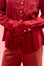 A close-up of a person wearing a dark red long-sleeve button-up shirt with gathered cuffs and matching dark red pants, set against a solid pink background.