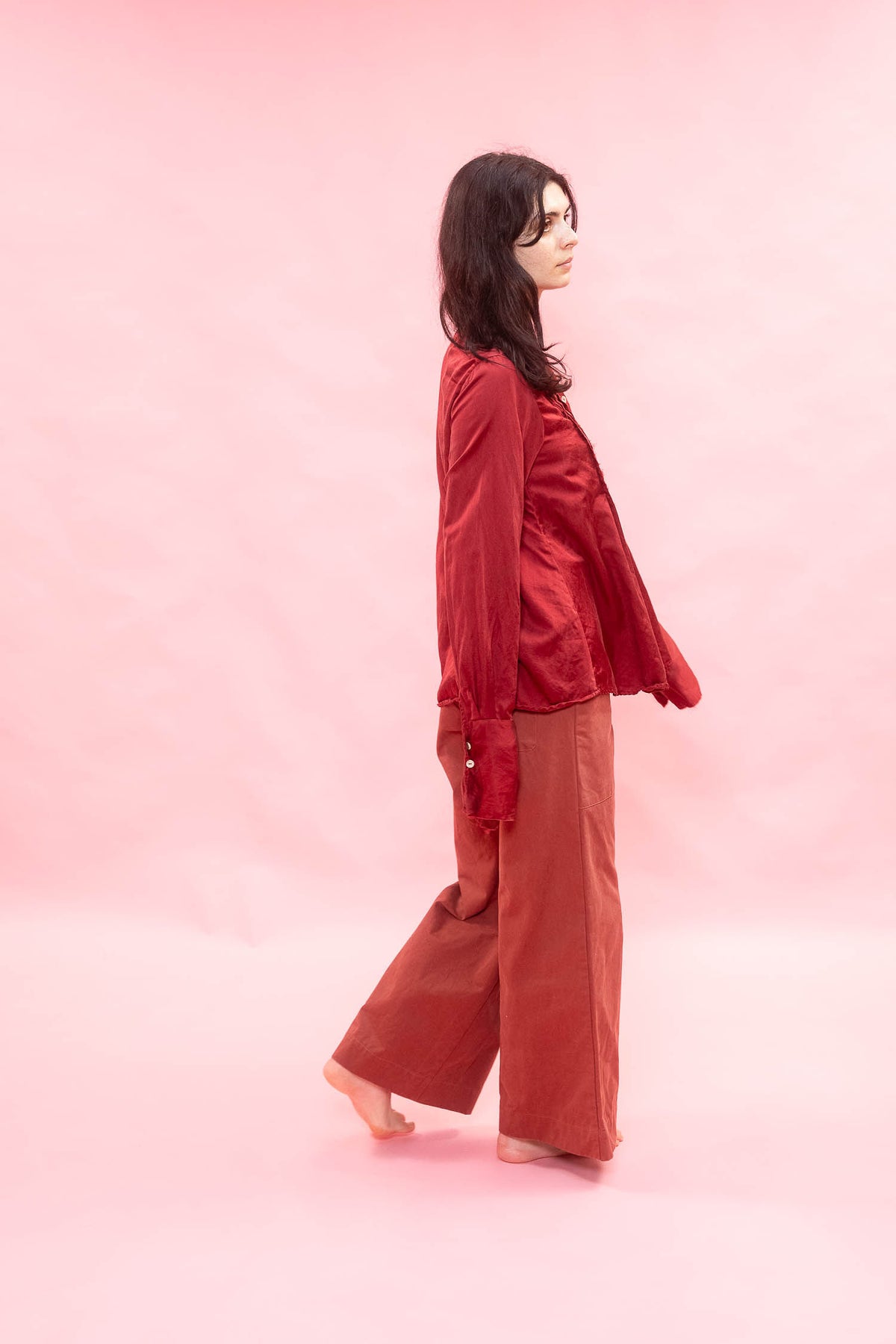 A woman stands barefoot in profile against a solid pink studio background, wearing a shiny dark red satin long-sleeve top and rust-colored wide-leg pants.