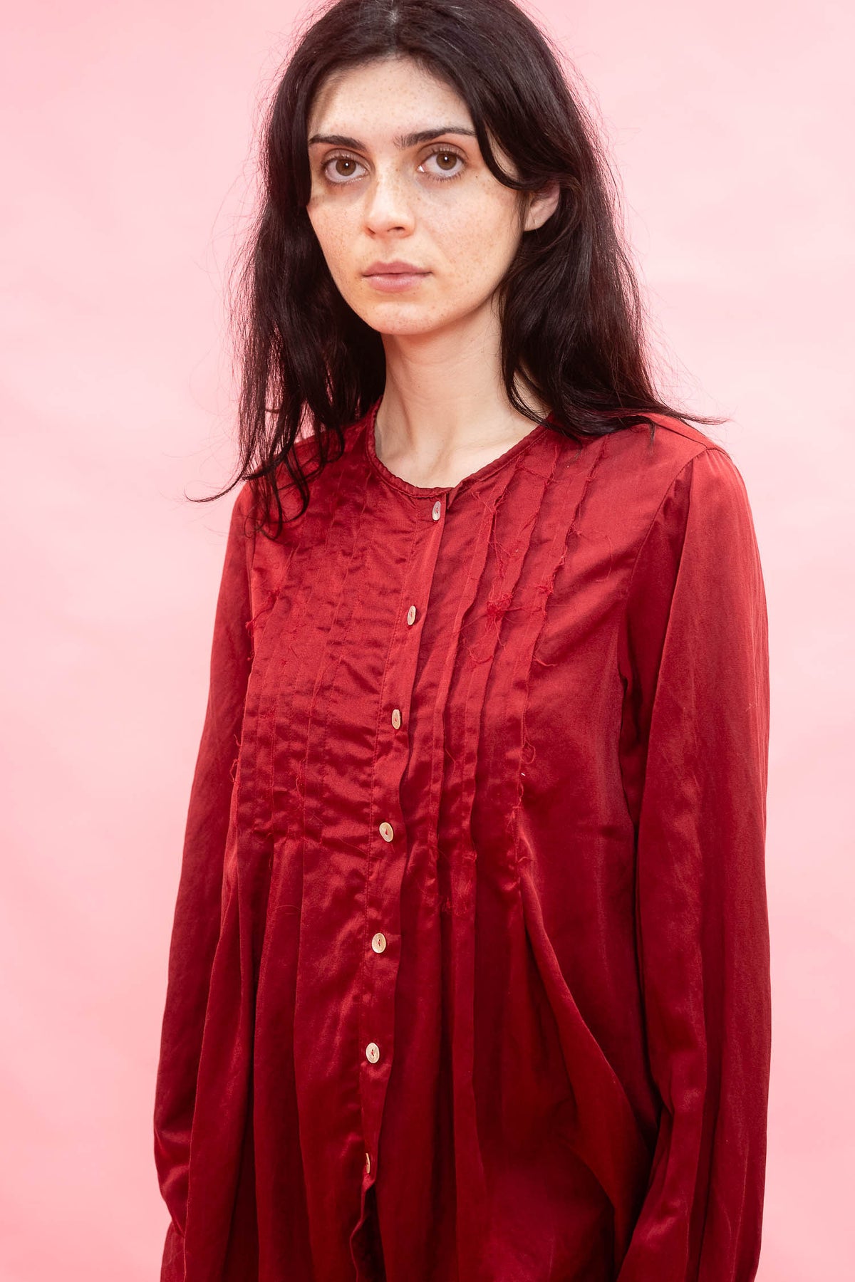 A woman with long dark hair wearing a textured, deep red long-sleeve button-up shirt, standing against a solid pink background.