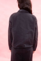 A person shown from the back wearing a dark charcoal or black cropped jacket with long sleeves, paired with matching dark pants, set against a solid pink background.
