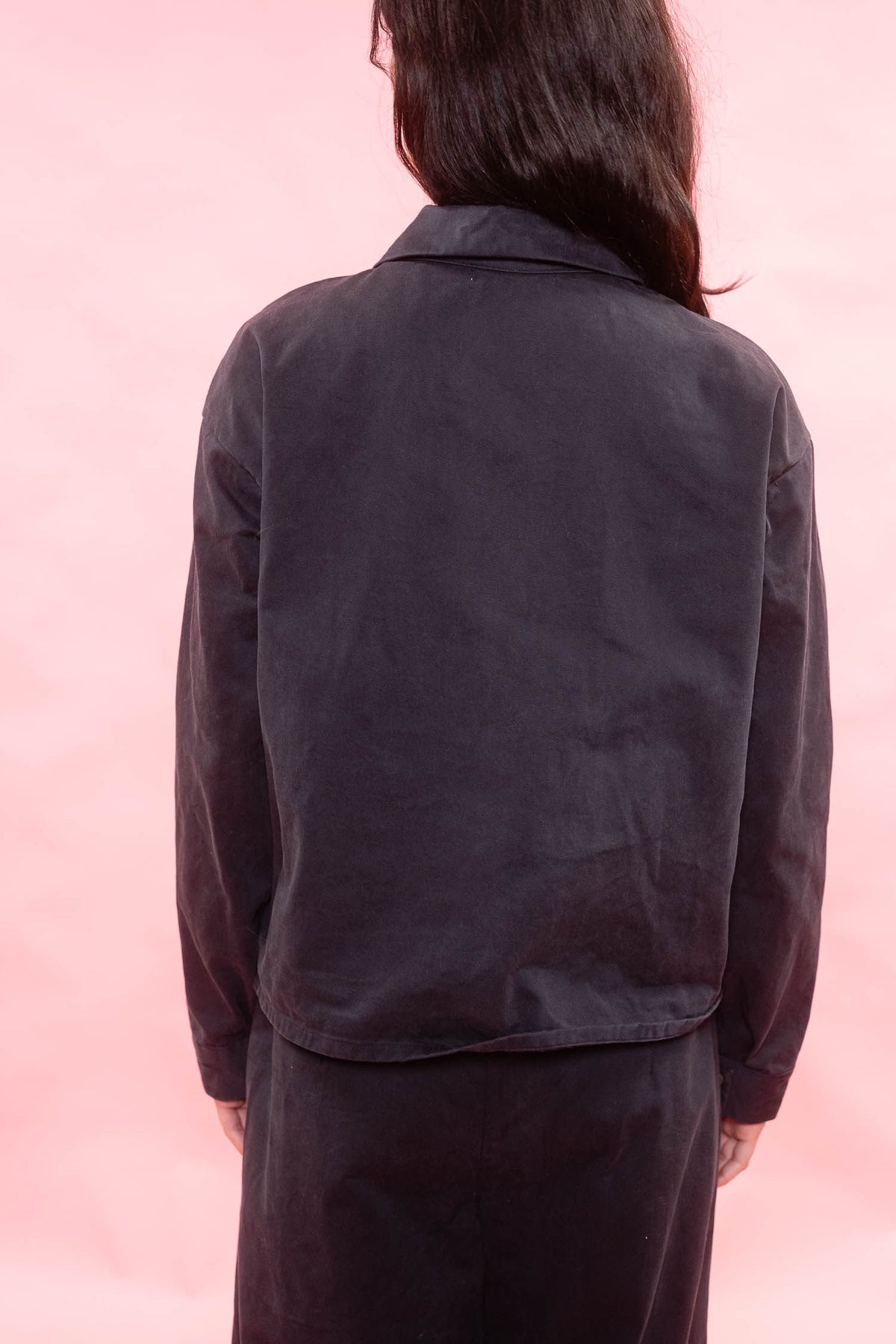 A person shown from the back wearing a dark charcoal or black cropped jacket with long sleeves, paired with matching dark pants, set against a solid pink background.