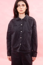A model wearing a black button-up shirt jacket with a chest pocket, photographed against a solid pink background.