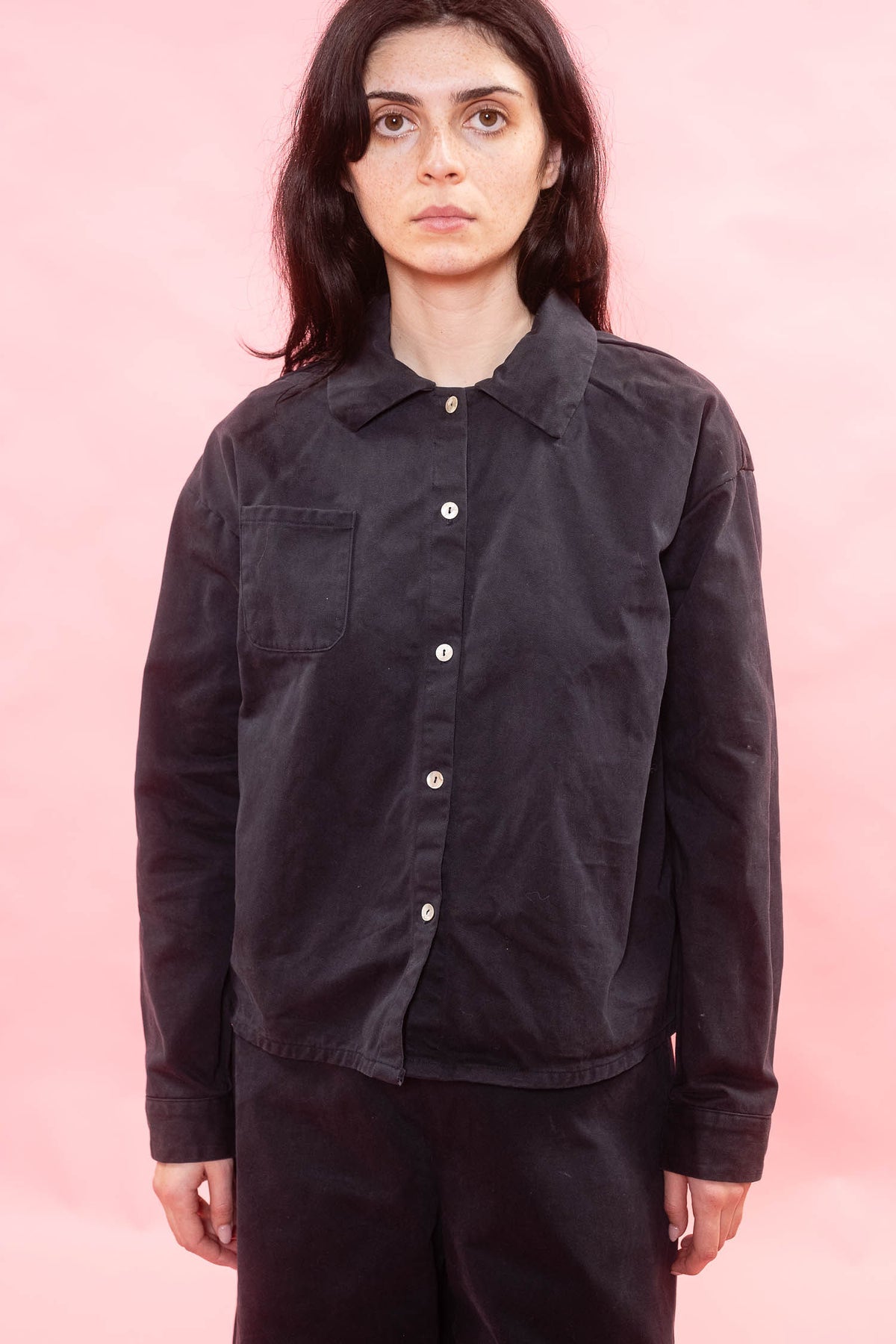 A model wearing a black button-up shirt jacket with a chest pocket, photographed against a solid pink background.