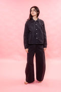 A model stands barefoot against a solid pink studio backdrop, wearing a black button-up shirt and matching black wide-leg pants.