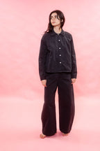A model stands barefoot against a solid pink studio backdrop, wearing a black button-up shirt and matching black wide-leg pants.