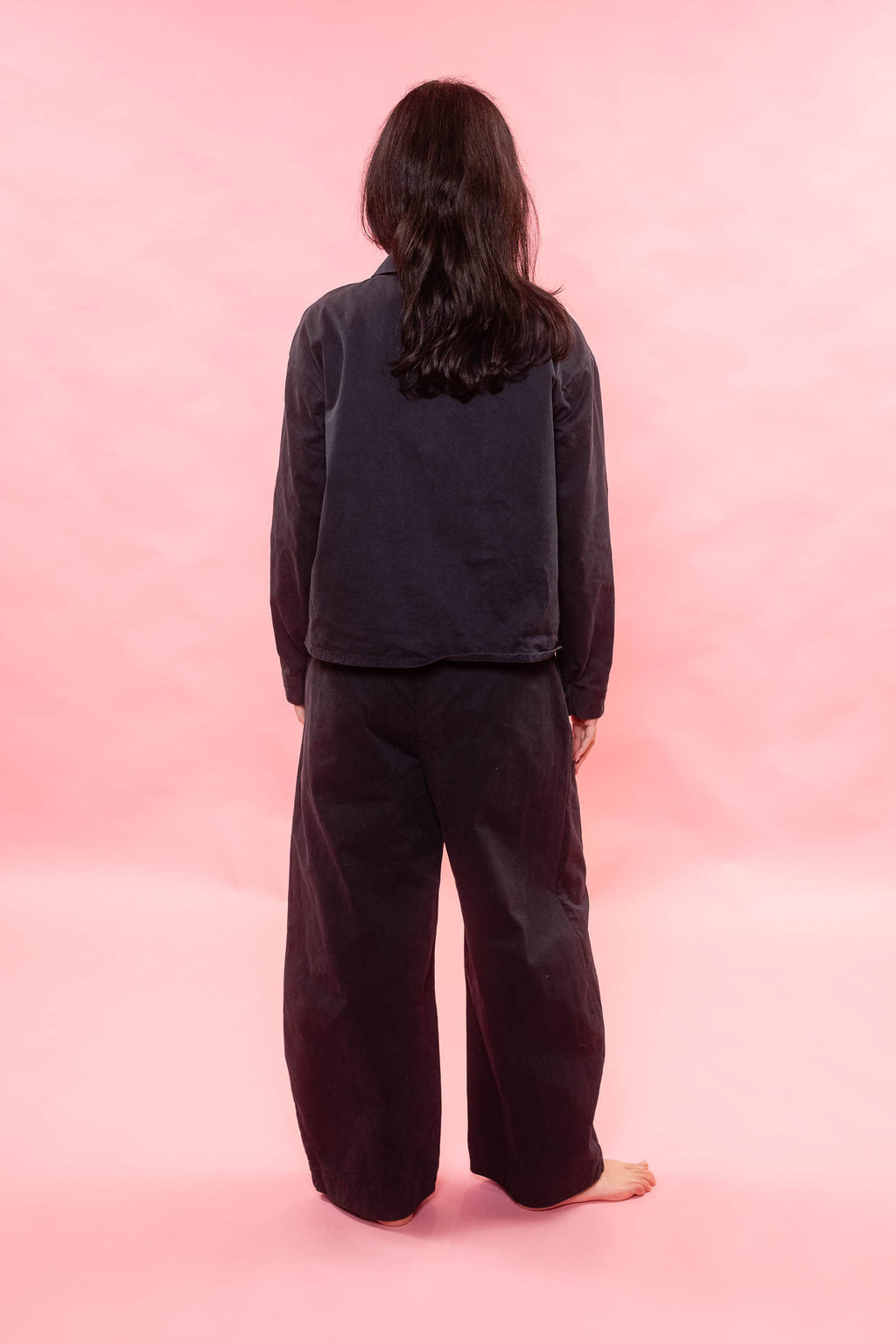 A person standing with their back to the camera, wearing a dark charcoal gray long-sleeve top and matching wide-leg pants against a solid pink studio background.