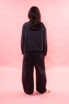A person standing with their back to the camera, wearing a dark charcoal gray long-sleeve top and matching wide-leg pants against a solid pink studio background.