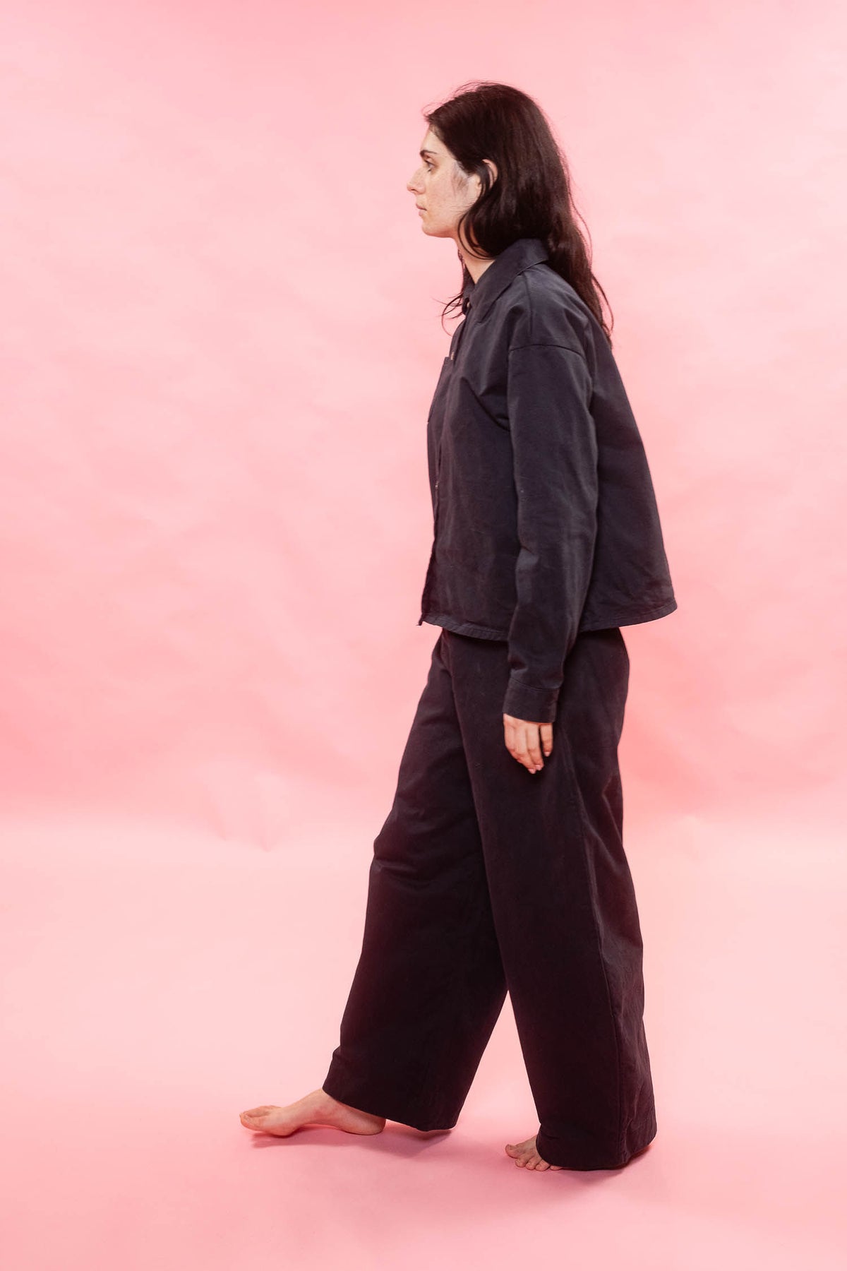 A woman stands in profile against a solid pink studio background, wearing a dark charcoal jacket and matching wide-leg pants.