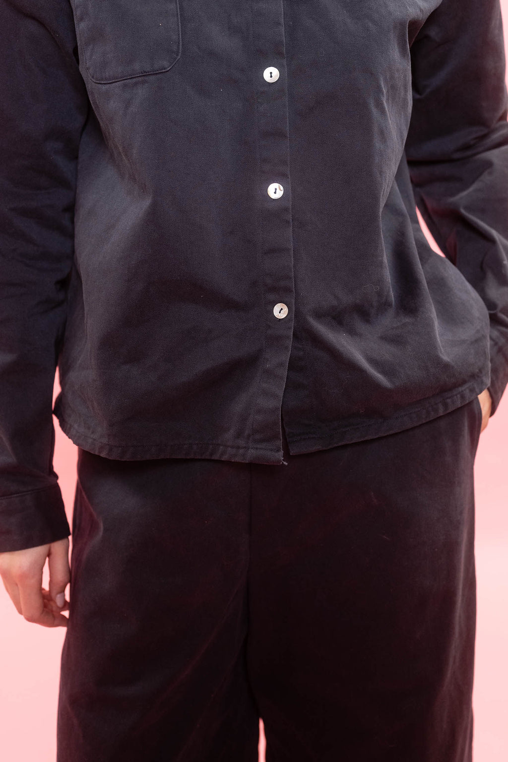 A close-up of a person wearing a dark charcoal button-up shirt and matching dark pants against a solid pink background.