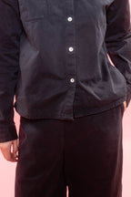 A close-up of a person wearing a dark charcoal button-up shirt and matching dark pants against a solid pink background.