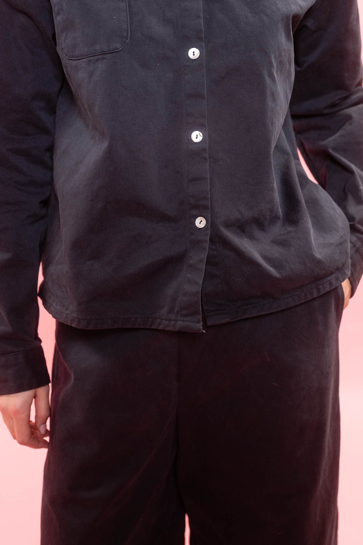 A close-up of a person wearing a dark charcoal button-up shirt and matching dark pants against a solid pink background.
