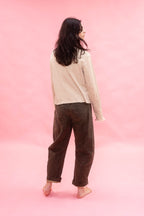 A woman is shown from behind wearing a light beige long-sleeve shirt and dirt green cotton patina pants, standing barefoot against a solid pink studio backdrop.