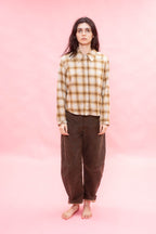 A woman stands barefoot against a solid pink studio background, wearing a long-sleeve button-up shirt with an ochre and brown plaid pattern made from Japanese soft brushed cotton wool mix, and loose dark brown pants.
