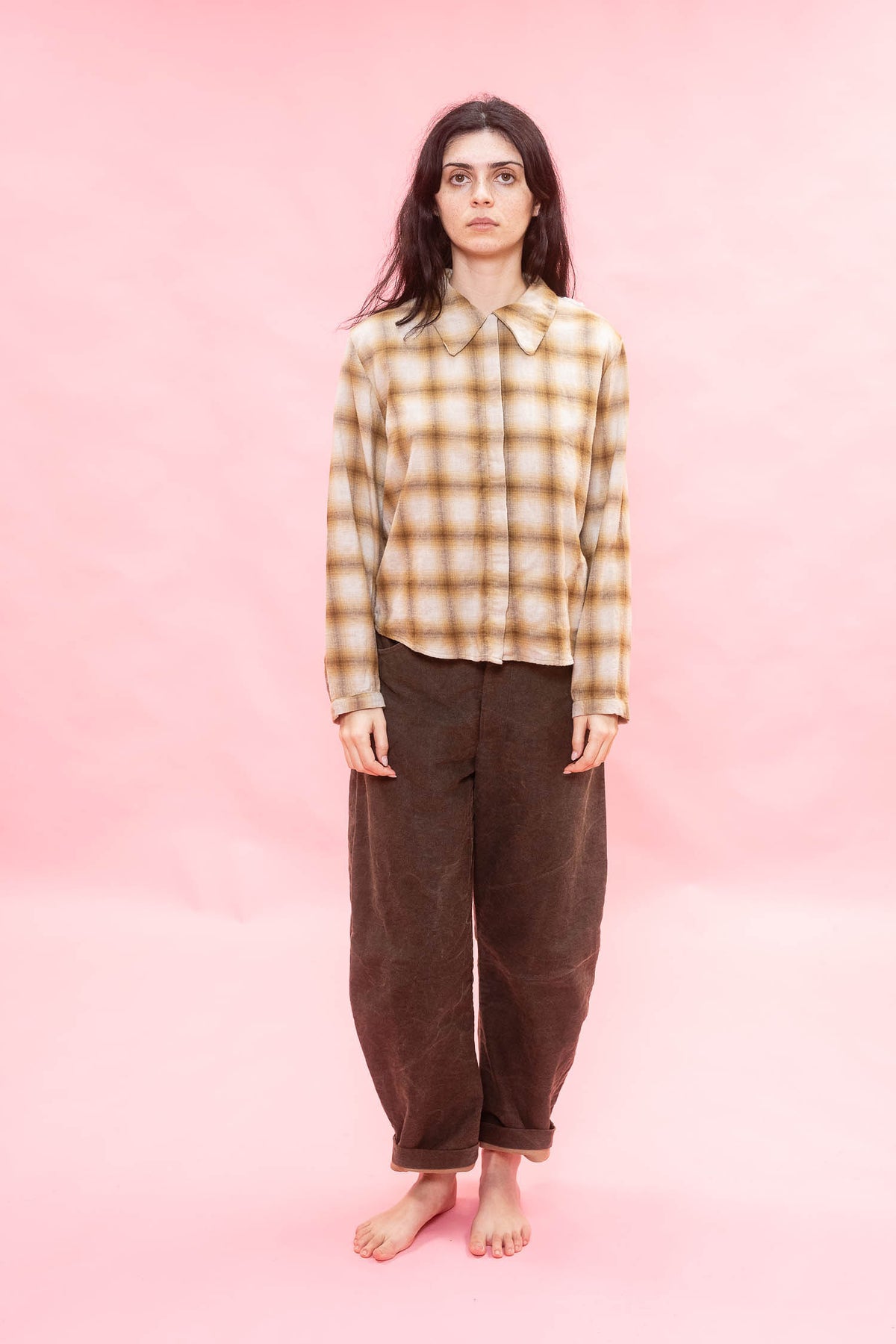 A woman stands barefoot against a solid pink studio background, wearing a long-sleeve button-up shirt with an ochre and brown plaid pattern made from Japanese soft brushed cotton wool mix, and loose dark brown pants.

