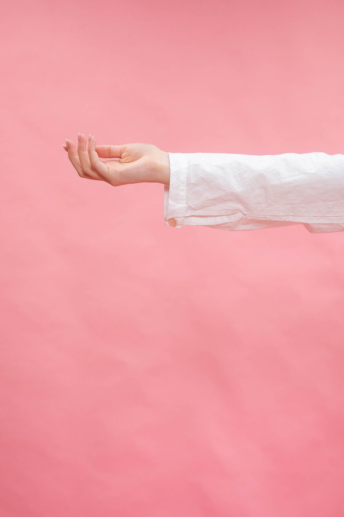 A person's hand and forearm wearing a white long-sleeve shirt, posed against a solid pink background.
