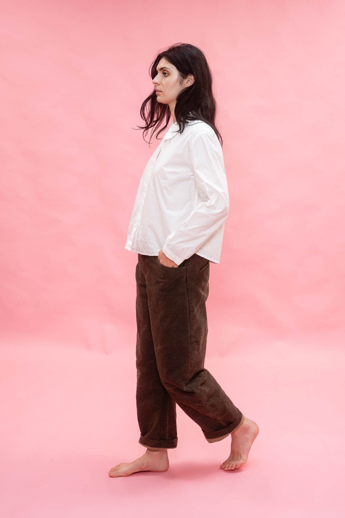 A person standing barefoot against a solid pink studio backdrop, wearing an oversized white long-sleeve shirt and loose dark brown pants.