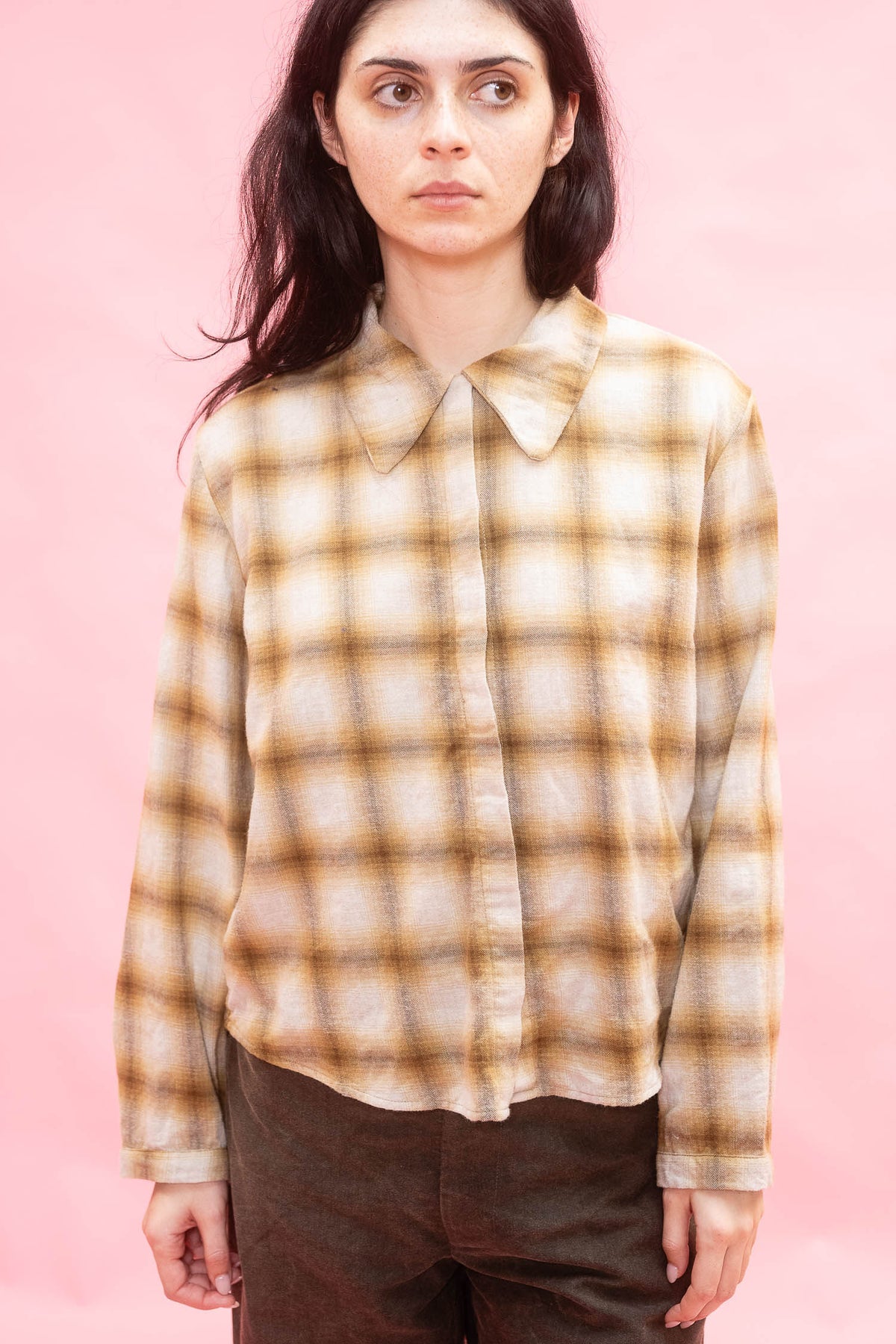 A woman wearing a cream and brown plaid button-down shirt with a pointed collar,  made with Japanese soft brushed cotton wool mix in ochre paired with dark brown pants, set against a solid pink background.
