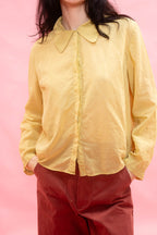 A person wearing a pale yellow long-sleeve button-front shirt with a pointed collar, paired with rust red pants, against a solid pink background.