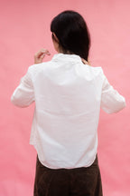 A person viewed from behind wearing a white long-sleeve button-up shirt and dark brown pants, standing against a solid pink background.