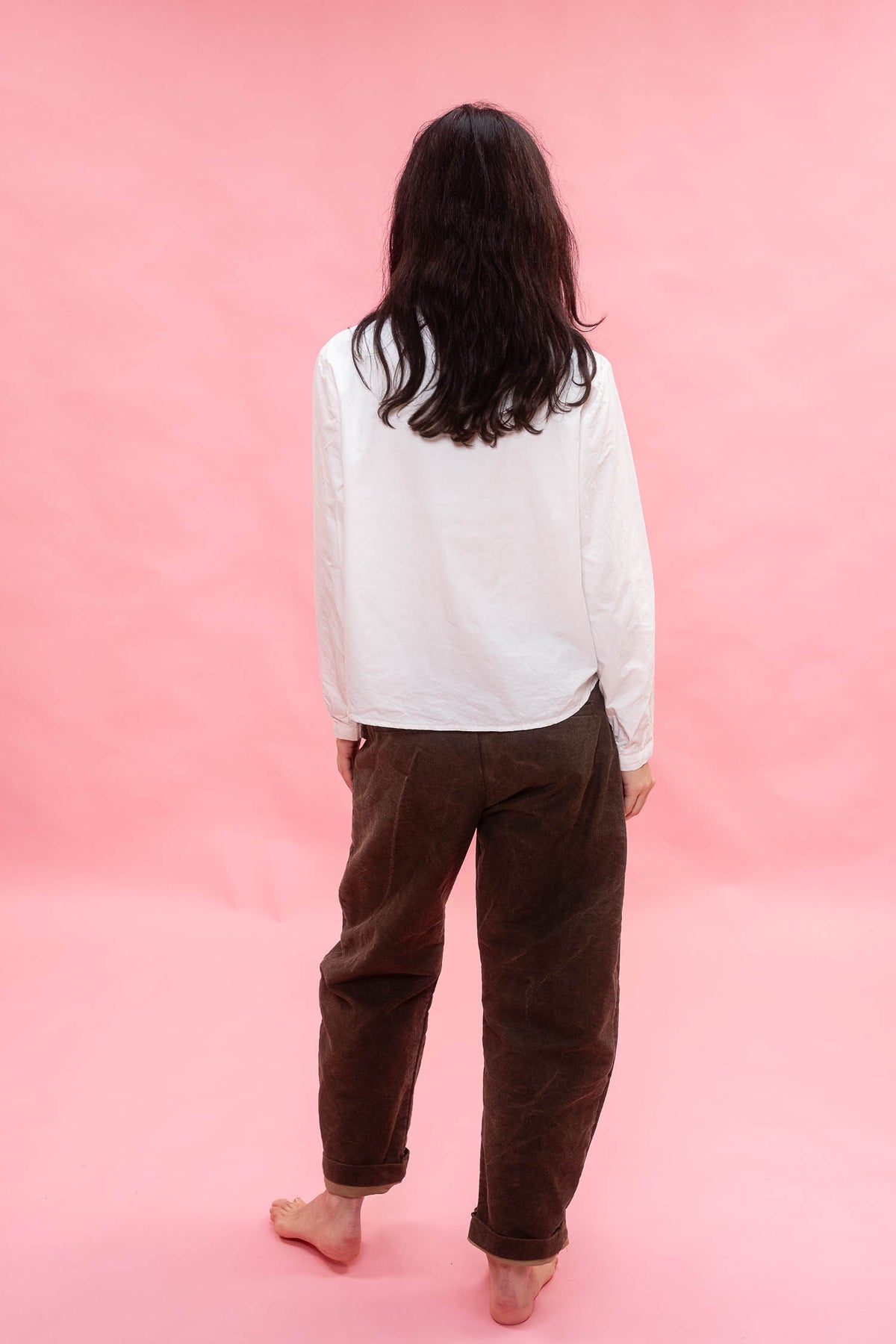 A person standing with their back to the camera, wearing a white long-sleeve shirt and dark brown corduroy pants, standing barefoot against a solid pink background.