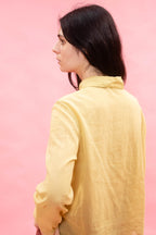 A woman with dark hair is shown from the back wearing a pale yellow linen shirt against a solid pink background.