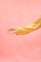 A close-up of a person's hand and forearm wearing a yellow button-up shirt, set against a solid pink background.