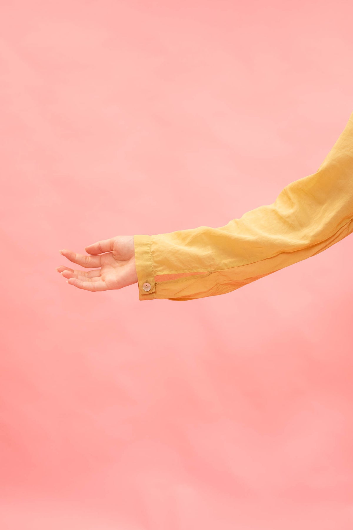 A close-up of a person's hand and forearm wearing a yellow button-up shirt, set against a solid pink background.