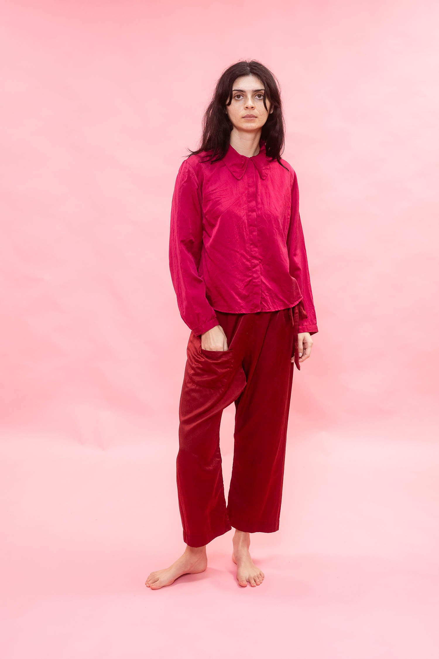 A woman stands barefoot against a solid pink studio background, wearing a shiny magenta long-sleeve button-up shirt and deep red wide-leg pants.