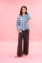 A woman stands barefoot against a solid pink studio backdrop, wearing a blue and white plaid button-up shirt and dark navy wide-leg pants.