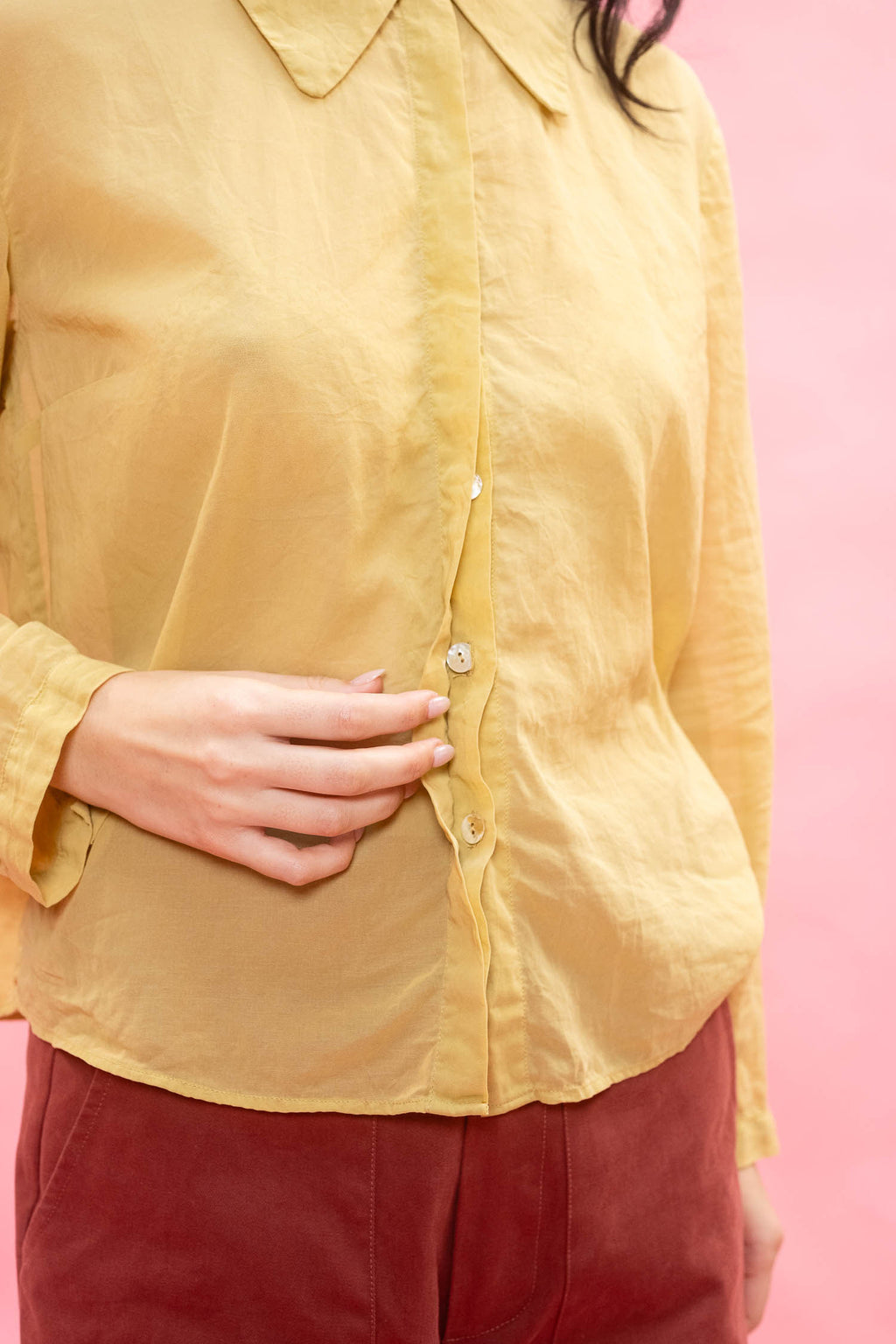 A close-up of a person wearing a mustard yellow linen button-up shirt and burgundy pants against a solid pink background.
