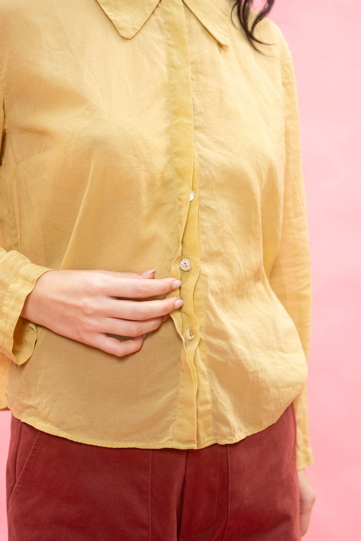 A close-up of a person wearing a mustard yellow linen button-up shirt and burgundy pants against a solid pink background.