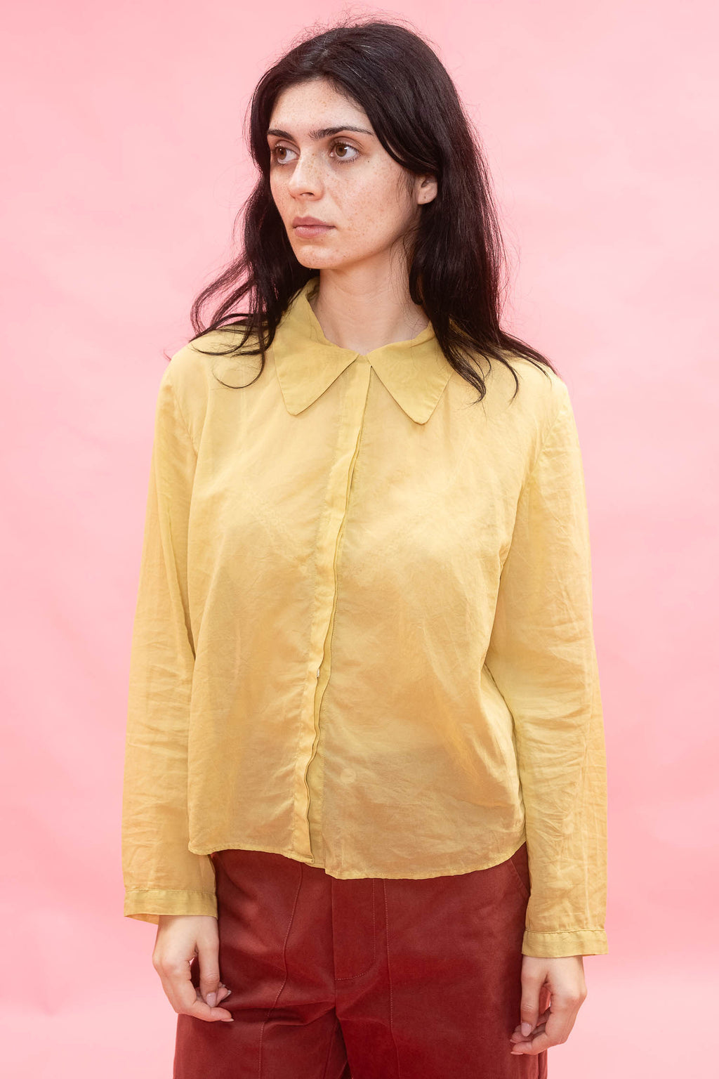 A woman wearing a pale yellow long-sleeve collared blouse and dark red trousers, standing against a solid pink studio background.