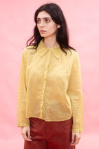 A woman wearing a pale yellow long-sleeve collared blouse and dark red trousers, standing against a solid pink studio background.