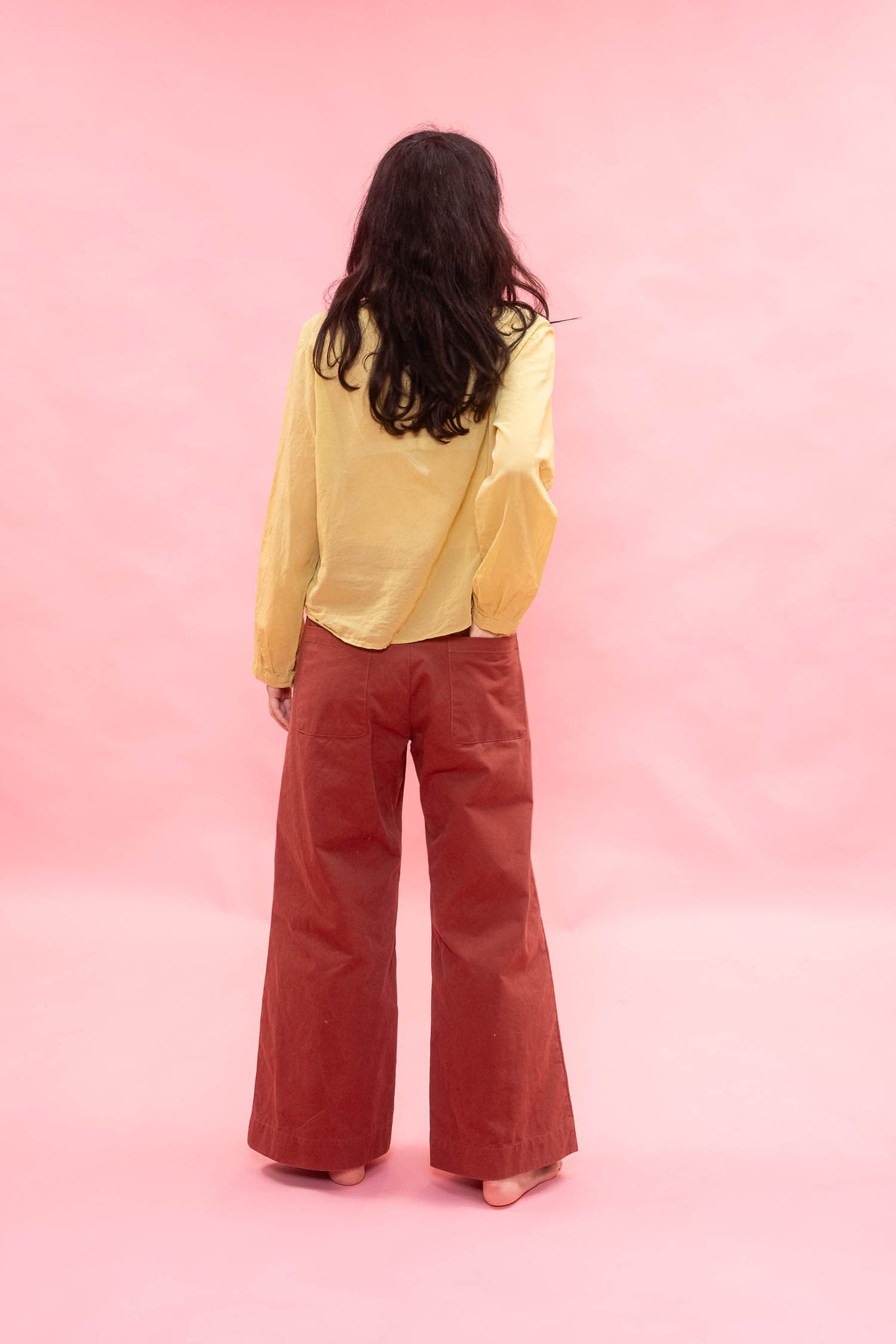 A woman is shown from behind wearing a yellow long-sleeve blouse and rust-colored wide-leg pants, standing against a solid pink studio backdrop.