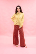 A woman stands barefoot against a solid pink studio backdrop, wearing a pale yellow long-sleeve button-up shirt and rust-colored wide-leg pants.