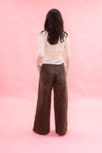 A woman viewed from behind wearing a white long-sleeve sheer top made from stretch knitted silk tulle, and dark brown wide-leg  pants, standing against a solid pink studio backdrop.
