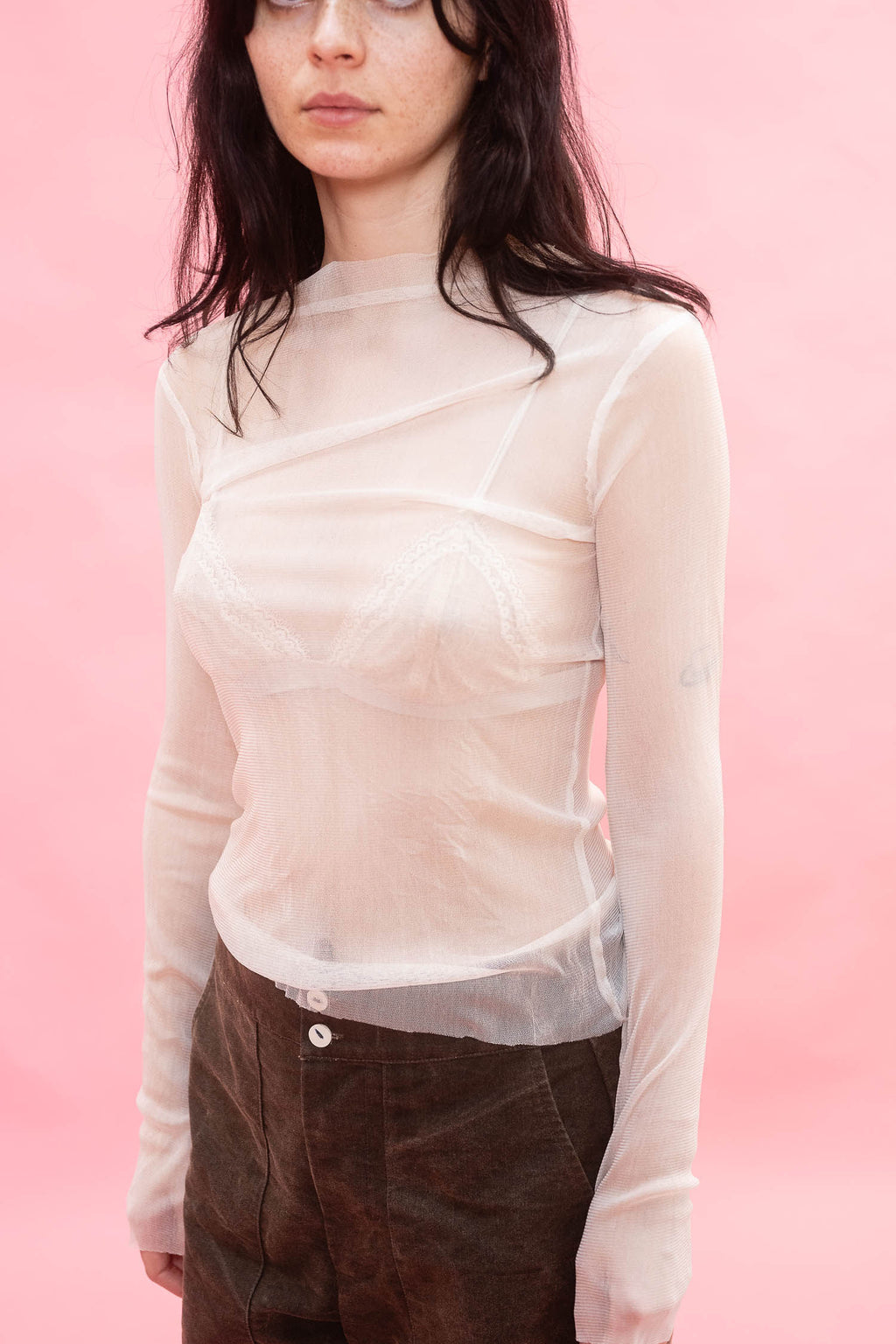 A model wearing a sheer, semi-transparent long-sleeve top with a structured cut, made from stretch knitted silk tulle, paired with brown pants, set against a solid pink background.
