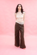 A woman stands against a solid pink studio backdrop wearing a sheer cream long-sleeve top made from stretch knitted silk tulle,and dark brown wide-leg corduroy pants.
